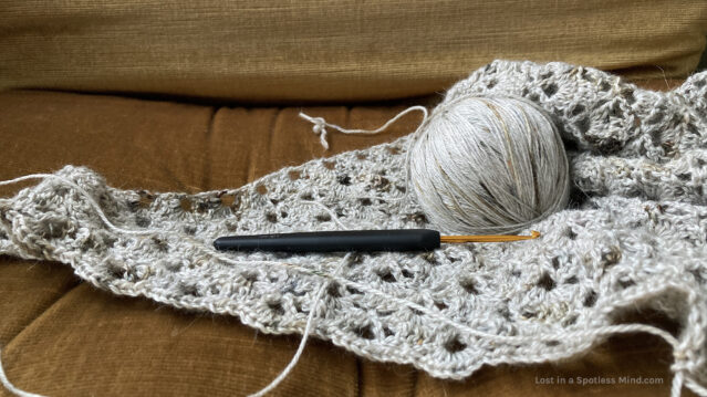 A photo of a lacy, light grey crochet shawl in progress