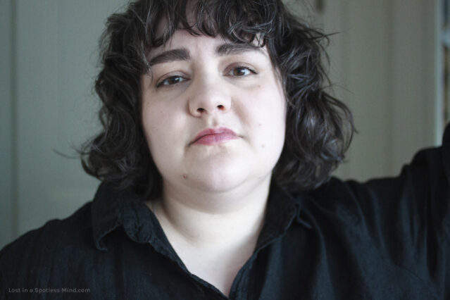 A headshot of Maria, a white woman in her 30s. She has dark, wavy hair and a fringe, brown eyes, and is wearing a black shirt. Her head is tilted up and back, and she looks straight into the camera.