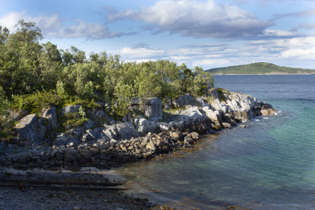 A seascape, where rocks and trees and a tiny boathouse jut out into a blue-green sea.
